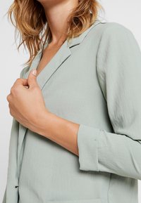 Vaaleanvihreä, teksturoitu bleiseri, jossa notkoreunaiset kauluskäänteet ja käännetyt hihat; istuu kapeasti ja on valmistettu pehmeästä, laskeutuvasta kankaasta.