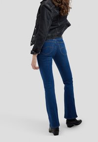 Femme portant un jean bleu foncé, des bottines noires et une veste en denim noire, debout légèrement tournée sur un fond uni clair.
