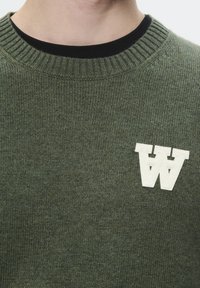 Grüner gestrickter Pullover mit einem gerippten Halsausschnitt und einem cremefarbenen "W"-Logo auf der linken Seite. Die Stofftextur wirkt weich und kuschelig.