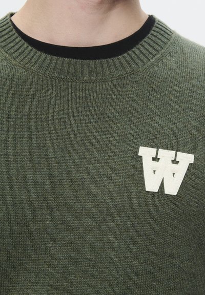 Pull en tricot vert avec un col côtelé et un logo "W" de couleur crème sur le côté gauche. La texture du tissu semble douce et confortable.