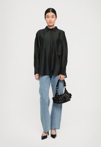 Kvinne iført løs svart skjorte med knapper, lyseblå rette jeans, svarte spisse hæler, og holder en svart naglet håndveske mot en hvit bakgrunn.