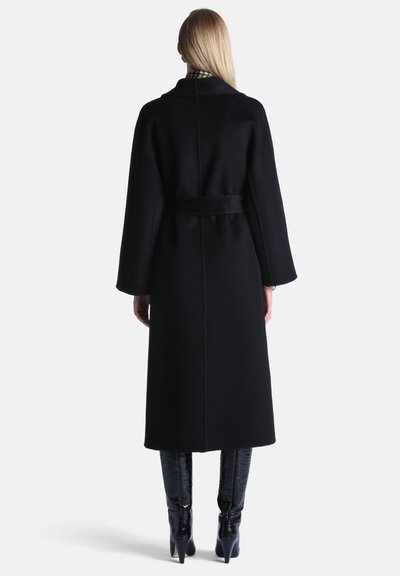 Femme portant un long manteau noir à manches larges, ceinturé à la taille, associé à des bottes noires texturées à talons montant jusqu'aux genoux, vue de dos.