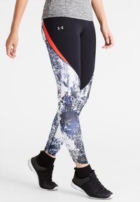 Ben med svart, vit och blåmönstrade Under Armour leggings med en röd rand och svarta träningsskor på en vit bakgrund.