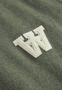 Grüner Strickstoff mit einem cremefarbenen gestickten "W"-Logo. Die Textur ist weich mit einer subtilen Farbvariation.