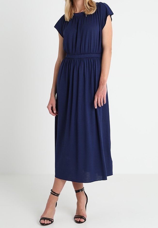 Robe bleu marine jusqu'aux genoux, taille froncée, manches courtes et tissu lisse. Portée avec des talons noirs à brides.