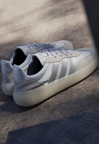Paire de baskets blanches Adidas avec des accents en daim gris et des semelles claires posées sur une surface en béton texturé sous une lumière douce du jour.