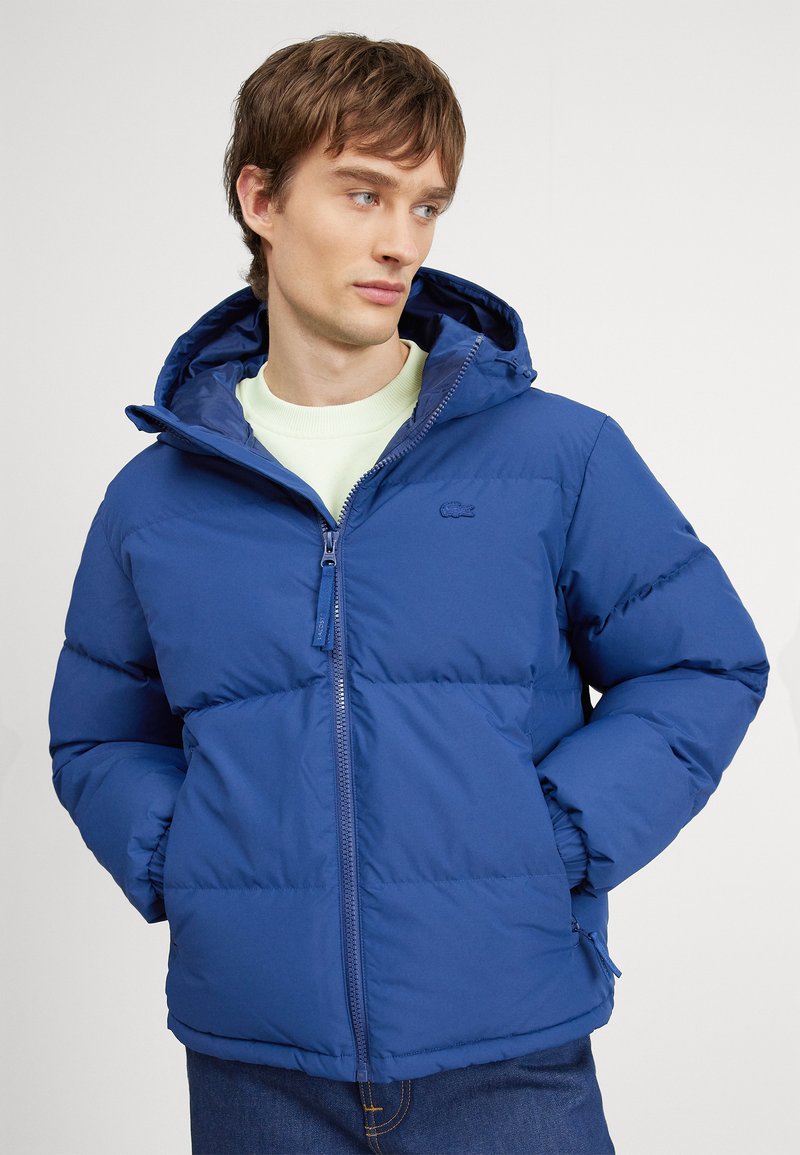 Joven vestido con una chaqueta acolchada azul, con las manos en los bolsillos, mirando hacia un lado contra un fondo blanco liso.