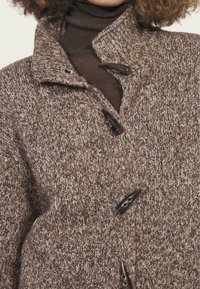 Hnědý pletený kabát s texturovaným povrchem, vybavený vysokým límcem a černými kovovými knoflíky pro zapínání. Materiál vypadá měkký a teplý.