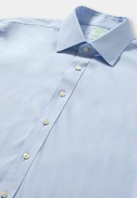 Lichtblauw overhemd met knopen, gemaakt van een gladde stof. Heeft een puntige kraag en zes witte knopen langs de voorlijst.