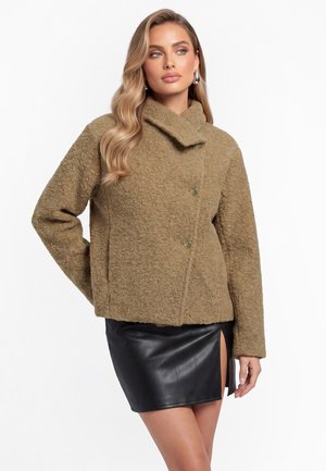 Frau mit langen blonden Haaren, die eine strukturierte braune Jacke und einen schwarzen Leder-Minirock mit Frontschlitz trägt, posiert vor einem weißen Hintergrund.
