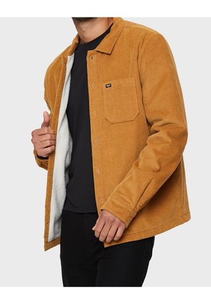 Hombre que lleva una chaqueta de pana de color mostaza con forro de forro polar blanco, mostrando el interior de la chaqueta mientras lleva una camisa negra y pantalones.