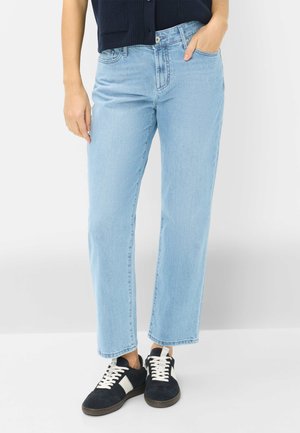 Person trägt hellblaue Jeans mit geradem Bein und Hand in der Tasche, schwarz-weiße Turnschuhe und ein dunkelblaues ärmelloses Oberteil.