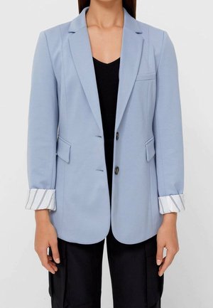 Kvinde iført en lyseblå blazer med stribede manchetter foldet tilbage, sort top og sorte bukser, stående mod en ensfarvet baggrund.