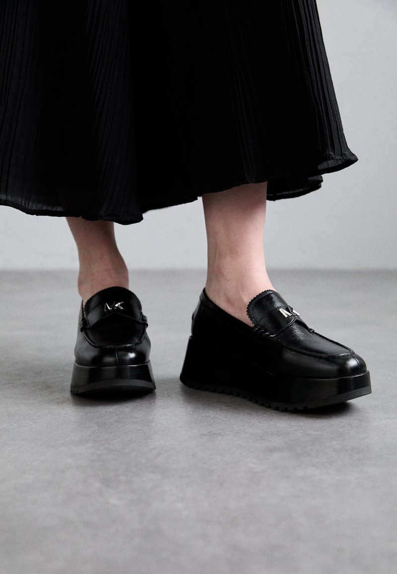 Mocassini neri con platform, finitura lucida, tomaia in pelle testurizzata e dettaglio hardware decorativo. Design slip-on con suola spessa.
