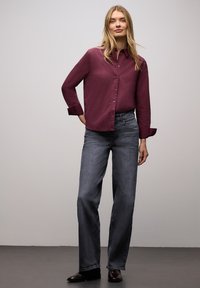 Bordowa koszula na guziki z długimi rękawami, w zestawieniu z jasnoszarymi szerokimi dżinsami. Model stoi na szarej podłodze, buty są ciemnobrązowe.