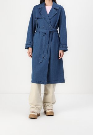 Femme portant un trench-coat en denim bleu ceinturé sur un pantalon beige clair avec des bottes camel, debout devant un fond blanc uni.