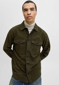 Mand iført olivengrøn skjorte med knapper over en beige turtleneck og blå jeans, stående med hænderne bag ryggen mod en helt hvid baggrund.