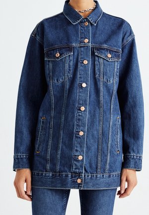 Veste en jean bleu foncé avec boutons en cuivre, poches poitrine à rabat, poches latérales fendues, portée avec un pantalon en jean assorti, et un collier chaîne argenté.