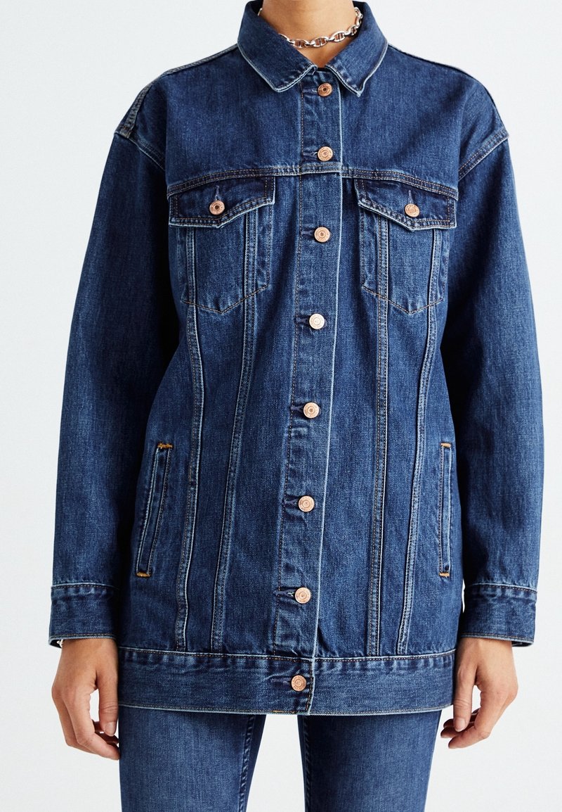Veste en jean bleu foncé avec boutons en cuivre, poches poitrine à rabat, poches latérales fendues, portée avec un pantalon en jean assorti, et un collier chaîne argenté.