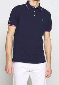 Námořnicky modré polokošile s límečkem a proužky na rukávech v červené a bílé barvě, s malým vyšívaným logem na hrudi.