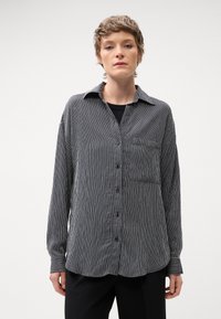 Camicia a righe bianche e nere con bottoni, maniche lunghe, colletto e taschino sul petto. Il tessuto ha una texture liscia e una vestibilità comoda.