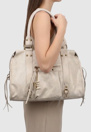 Donna che porta una grande borsa in pelle beige con tasca frontale con cerniera, borchie color oro e ciondolo con logo sulla spalla su sfondo semplice.
