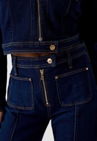 Close-up van een donkerblauwe spijkerjas en broek met gouden stiksels, voorzien van zichtbare ritsen en metalen knoopsluitingen.