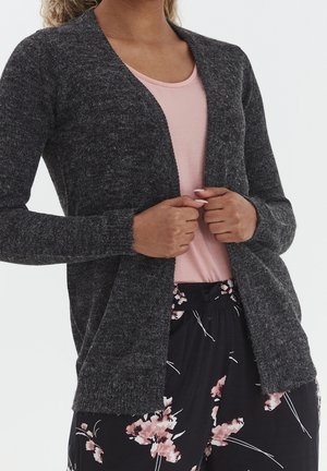 Kvinde iført en mørkegrå cardigan med åbent front over en lyserød top og sorte bukser med blomsterprint, der holder kardiganens kanter.