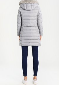 Polstret grå frakke med en faux fur hætte, lange ærmer, tætsiddende form og elastisk talje, parret med en mørk jeans og lyse sneakers.