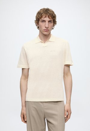 Ung mand med lyst krøllet hår iført en cremefarvet kortærmet polo og beige bukser, stående foran en ensfarvet hvid baggrund.