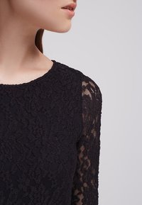 Svart spetsöverdel med rundad halsringning, texturerat blommönster på kroppen och genomskinliga spetslånga ärmar.