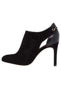 Botte à talons hauts en suède noir avec un design latéral ouvert, présentant un accent en simili cuir et un détail en chaîne dorée au niveau de la cheville.