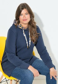 Námořnická modrá mikina s kapucí, která má vzorovanou vnitřní podšívku, šňůrky se zlatými a černými akcenty a volný střih. Nosené s modrými džínami.