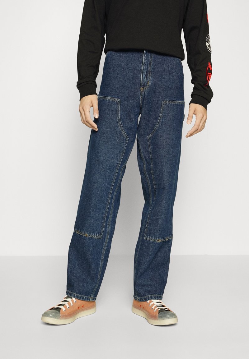 Jeans de mezclilla azul con un corte holgado, que presentan grandes bolsillos frontales y un diseño de pierna recta. Combinados con zapatillas bajas naranjas.