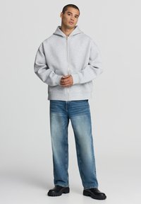 Giovane uomo in piedi che indossa un felpa chiusa con zip grigia chiara, jeans blu sbiaditi e stivali neri robusti, davanti a uno sfondo uniforme chiaro.