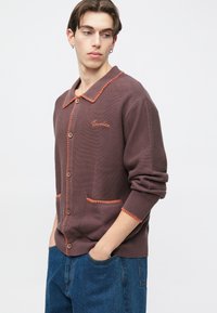 Cardigan na zapínanie na gombíky v tmavohnedej farbe s kontrastným oranžovým prešívaním, golierom a vreckovými detailmi. Mäkká textúra, voľný strih.