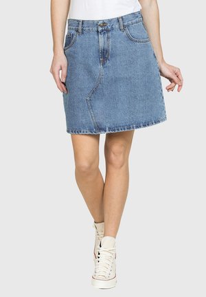 Donna che indossa una gonna di jeans blu al ginocchio con tasche frontali, sneakers bianche alte e un top bianco su sfondo semplice.