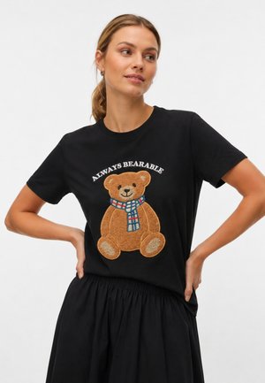 Donna che indossa una t-shirt nera con una grafica di un orsacchiotto e il testo "Always Bearable,", abbinata a una gonna nera, in piedi con le mani sui fianchi.