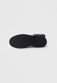 Semelle en caoutchouc noir d'une chaussure décontractée avec un motif de grip texturé, vue par le dessous sur un fond blanc uni.