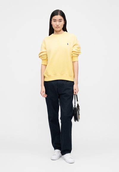 Yellow sweatshirt with a crew neckline and rolled sleeves, paired with dark blue jeans and white sneakers. The sweatshirt features a small logo detail.