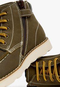Stivali ankle in pelle marrone con lacci gialli, zip laterale, cuciture bianche e suole in gomma. Presentano una finitura texturizzata e un collo allungato di forma distintiva.