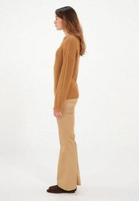 Pull en maille beige avec une texture côtelée, manches longues et détails en œillets, associé à un pantalon évasé marron clair et des chaussures marron foncé.