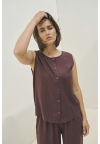 Bordový top bez rukávů s předním zapínáním na knoflíky a zaobleným lemem, vyrobený z jemné látky. Modelka je zkombinována s odpovídajícími kalhotami a jednoduchým náhrdelníkem.