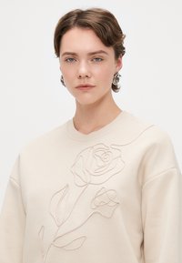 Μπεζ φούτερ με διακριτό σχέδιο κεντημένου τριαντάφυλλου. Το ύφασμα φαίνεται μαλακό με άνετη εφαρμογή και στρογγυλή λαιμόκοψη.