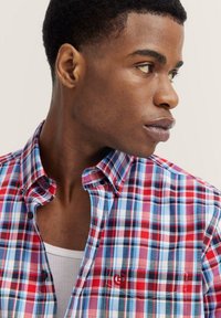 Karierte Baumwollbluse mit roten, blauen und weißen Karos, Knopfkragen und Logo-Akzent auf der Brust, mit einer glatten Textur.