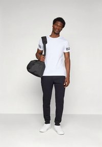 Bílá tričko s krátkým rukávem a černými detaily, doplněné tmavými tepláky a bílými teniskami. Černá taška přes rameno.