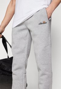 Grå joggebukser laget av myk stoff, med en sidelomme og et logo på forsiden. Designet er uformelt og smalner inn ved anklene.