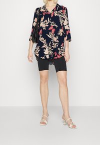 Kwiatowa tunika na granatowym tle, z rękawami o długości trzech ćwiartek i luźnym kroju; zestawiona z czarnymi szortami rowerowymi i białymi sandałkami na obcasie.