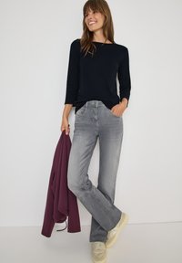 Top a maniche lunghe blu navy abbinato a jeans grigi a sigaretta. Il modello tiene in mano una giacca bordeaux, indossando sneakers beige. Design semplice, texture morbida.