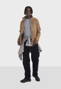 Braune Fleecejacke mit Reißverschlusstasche, graues T-Shirt, karierte Flanelljacke um die Taille gebunden, schwarze Cordhosen und schwarze Schuhe.
